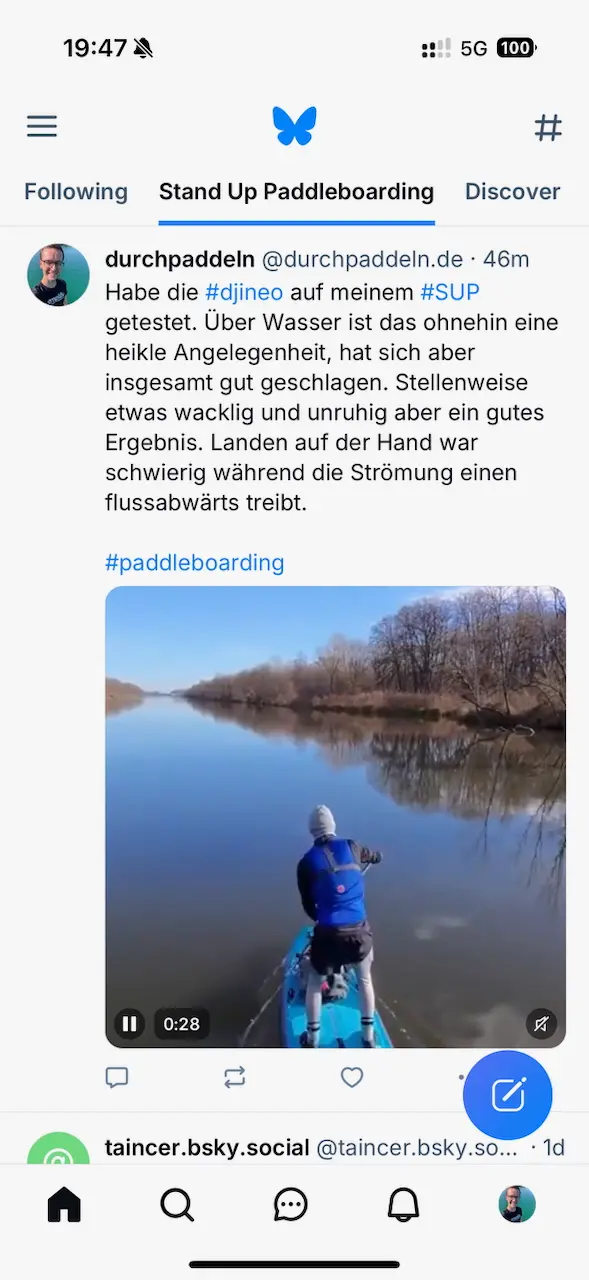 Screenshot der Bluesky-App im Feed "Stand Up Paddleboarding". Ein Beitrag von @durchpaddeln.de zeigt einen Text über das Testen einer Kamera (#djineo) auf einem Stand-Up-Paddleboard (#SUP). Der Beitrag beschreibt die Herausforderungen beim Filmen auf dem Wasser. Unter dem Text ist ein Video-Thumbnail zu sehen, das eine Person auf einem SUP zeigt, die einen ruhigen Fluss entlang paddelt. Im Hintergrund sind Uferbäume und ein klarer Himmel sichtbar. Oben befinden sich Menü- und Netzwerkstatussymbole, unten die Navigationsleiste der App.